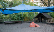 Spielplatz mit großem blauen Sonnenschirm, kleines Holzhaus und Sandkiste
