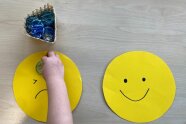 zwei Smileys - lachend und traurig - aus Pappe. Ein Kind legt Steine zur Bewertung drauf