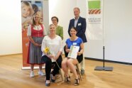 Mehrere erwachsene Personen stehen vor einer Wand, zwei Personen sitzen im Vordergrund und halten eine Urkunde in der Hand
