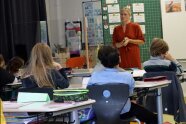 Frau steht in Klassenzimmer vor Kindern, die an Schultischen sitzen