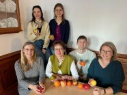 5 Frauen und ein Mann sitzen an einem Tisch. Alle halten sein Stück Obst in der Hand