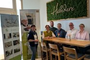6 Personden sitzen oder stehen an einem Tisch und lachen in die Kamera