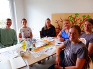5 Frauen und ein Mann sitzen am Tisch und lachen in die Kamera