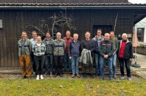 zwölf Personen sehen in die Kammera und stehe vor einem Schuppen auf einer Wiese