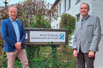 Zwei Männer stehen links und rechts neben einem Amtschild